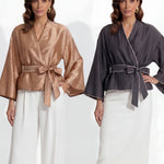 Two professional models wearing elegant shimmering satin wrap tops with kimono-style sleeves and waist ties, one in champagne gold and the other in charcoal grey with contrast white piping, paired with ivory high-waisted trousers.