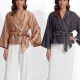 Two professional models wearing elegant shimmering satin wrap tops with kimono-style sleeves and waist ties, one in champagne gold and the other in charcoal grey with contrast white piping, paired with ivory high-waisted trousers.