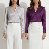 Two professional models wearing elegant V-neck silk satin blouses in shimmering silver and deep purple, featuring pleated front details and paired with white high-waisted trousers and a buttoned skirt.