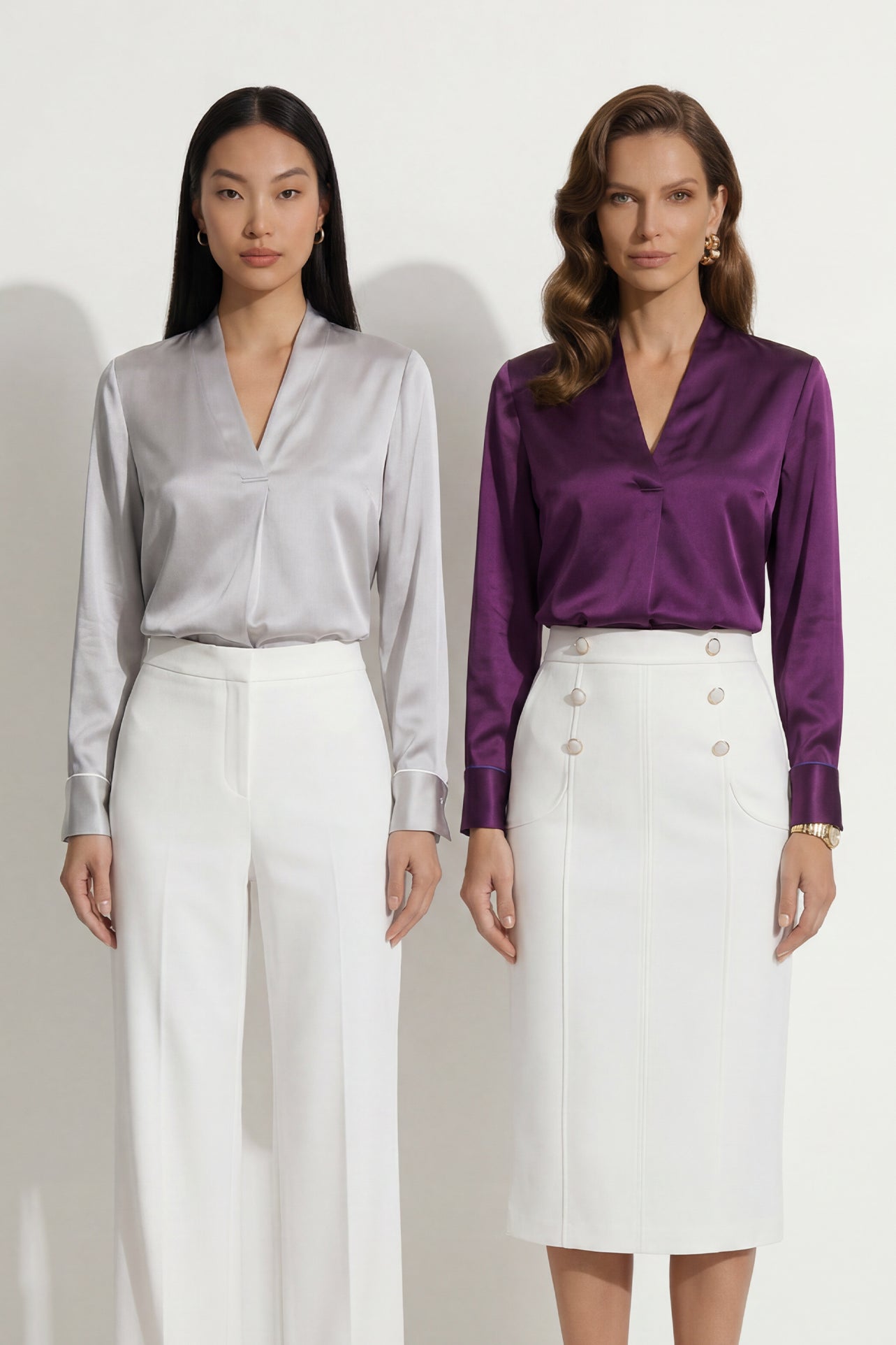 Two professional models wearing elegant V-neck silk satin blouses in shimmering silver and deep purple, featuring pleated front details and paired with white high-waisted trousers and a buttoned skirt.