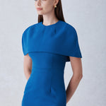 A professional woman wearing a sophisticated teal blue midi dress with an elegant cape-style overlay and structured shoulders, perfect for formal executive events and ceremonial occasions.