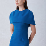 A professional woman wearing a sophisticated teal blue midi dress with an elegant cape-style overlay and structured shoulders, perfect for formal executive events and ceremonial occasions.
