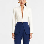 A professional model showcasing an elegant two-tone women's pantsuit, featuring a tailored cropped white blazer with a sharp deep V-neckline, paired with high-waisted navy blue trousers that have a unique curved overlay panel design and a golden metal hardware accent at the waist.