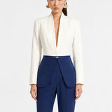 A professional model showcasing an elegant two-tone women's pantsuit, featuring a tailored cropped white blazer with a sharp deep V-neckline, paired with high-waisted navy blue trousers that have a unique curved overlay panel design and a golden metal hardware accent at the waist.