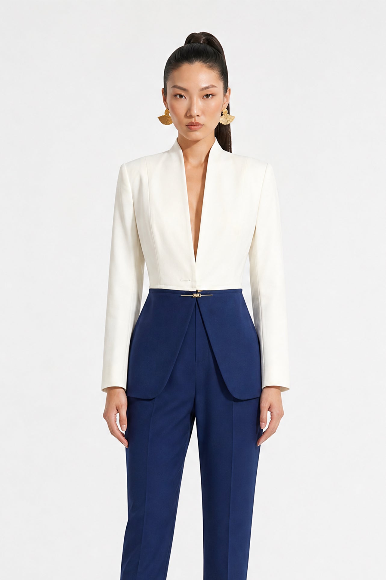 A professional model showcasing an elegant two-tone women's pantsuit, featuring a tailored cropped white blazer with a sharp deep V-neckline, paired with high-waisted navy blue trousers that have a unique curved overlay panel design and a golden metal hardware accent at the waist.