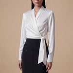 A professional woman wearing a high-end white silk wrap blouse with a side-tie detail, paired with a tailored black high-waisted pencil skirt for a sophisticated executive office look.