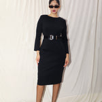 A professional woman wearing a sophisticated black long-sleeve sheath dress, featuring a classic crew neckline and a slim black belt with a gold buckle for a polished executive office look.