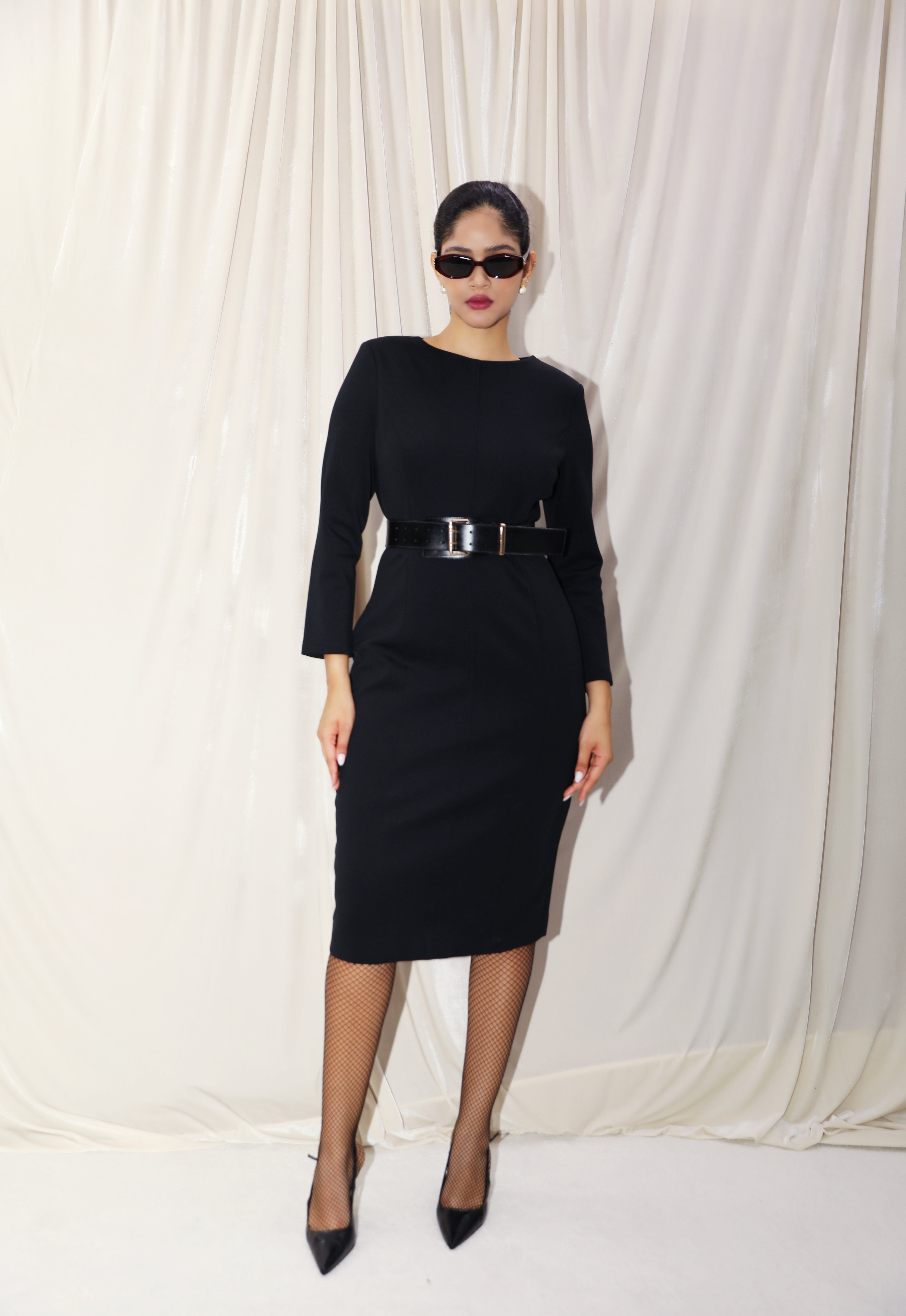 A professional woman wearing a sophisticated black long-sleeve sheath dress, featuring a classic crew neckline and a slim black belt with a gold buckle for a polished executive office look.