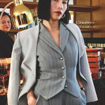 A professional woman wearing a sophisticated light grey tailored suit set, featuring a unique wrap-front blazer with criss-cross waist detailing and matching slim-fit trousers for a polished executive look.