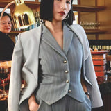A professional woman wearing a sophisticated light grey tailored suit set, featuring a unique wrap-front blazer with criss-cross waist detailing and matching slim-fit trousers for a polished executive look.