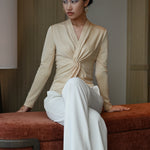 A chic Asian woman modeling an elegant champagne gold long-sleeve top featuring a sophisticated v-neckline and a stylized twist-front knot waist, beautifully paired with high-waisted white trousers.