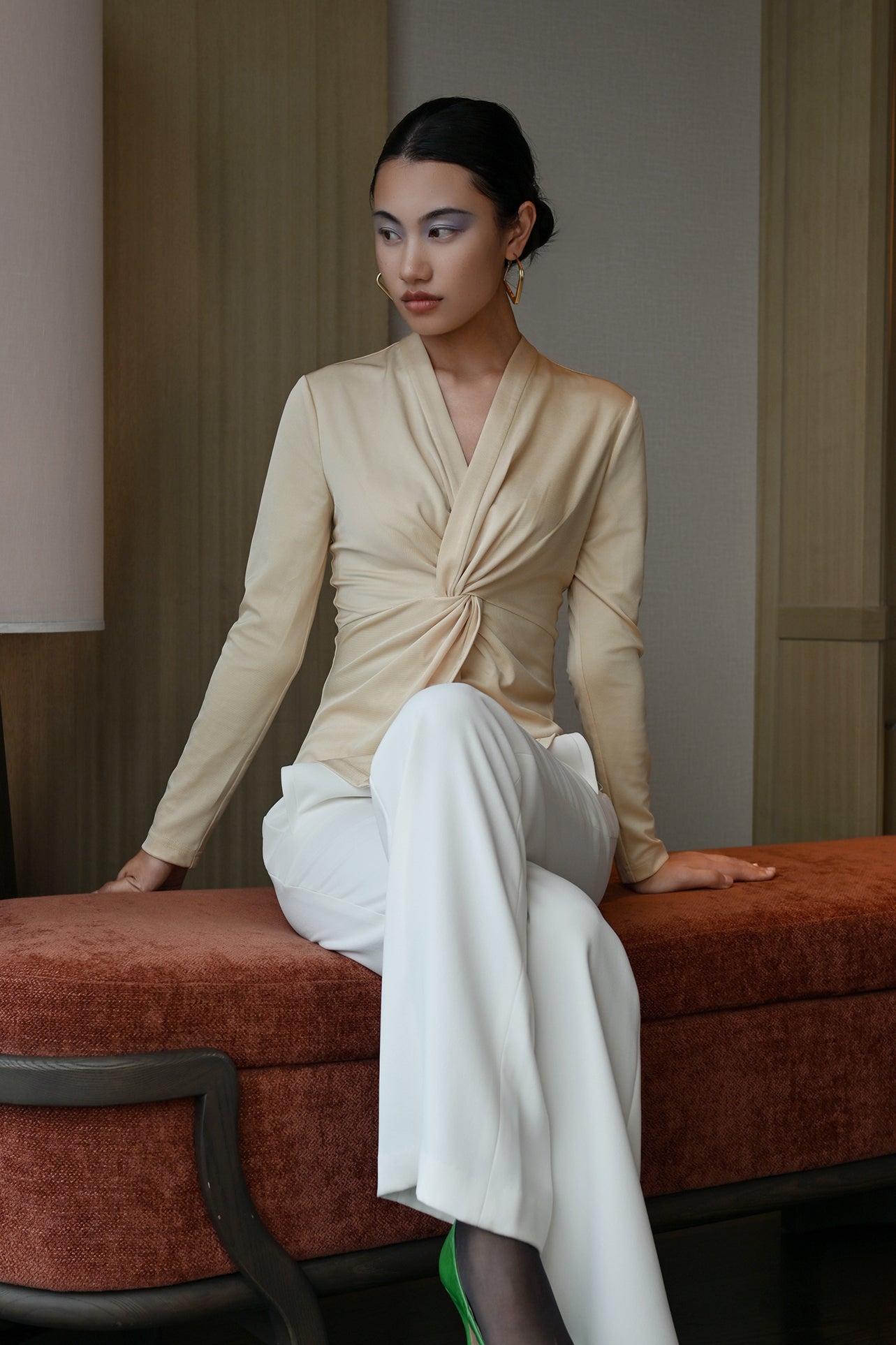 A chic Asian woman modeling an elegant champagne gold long-sleeve top featuring a sophisticated v-neckline and a stylized twist-front knot waist, beautifully paired with high-waisted white trousers.