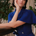An elegant woman wearing a sophisticated royal cobalt blue trench dress featuring a structured turn-down collar, a cropped capelet overlay, a double-breasted button front, and a cinched self-tie belted waist.