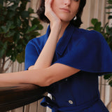 An elegant woman wearing a sophisticated royal cobalt blue trench dress featuring a structured turn-down collar, a cropped capelet overlay, a double-breasted button front, and a cinched self-tie belted waist.