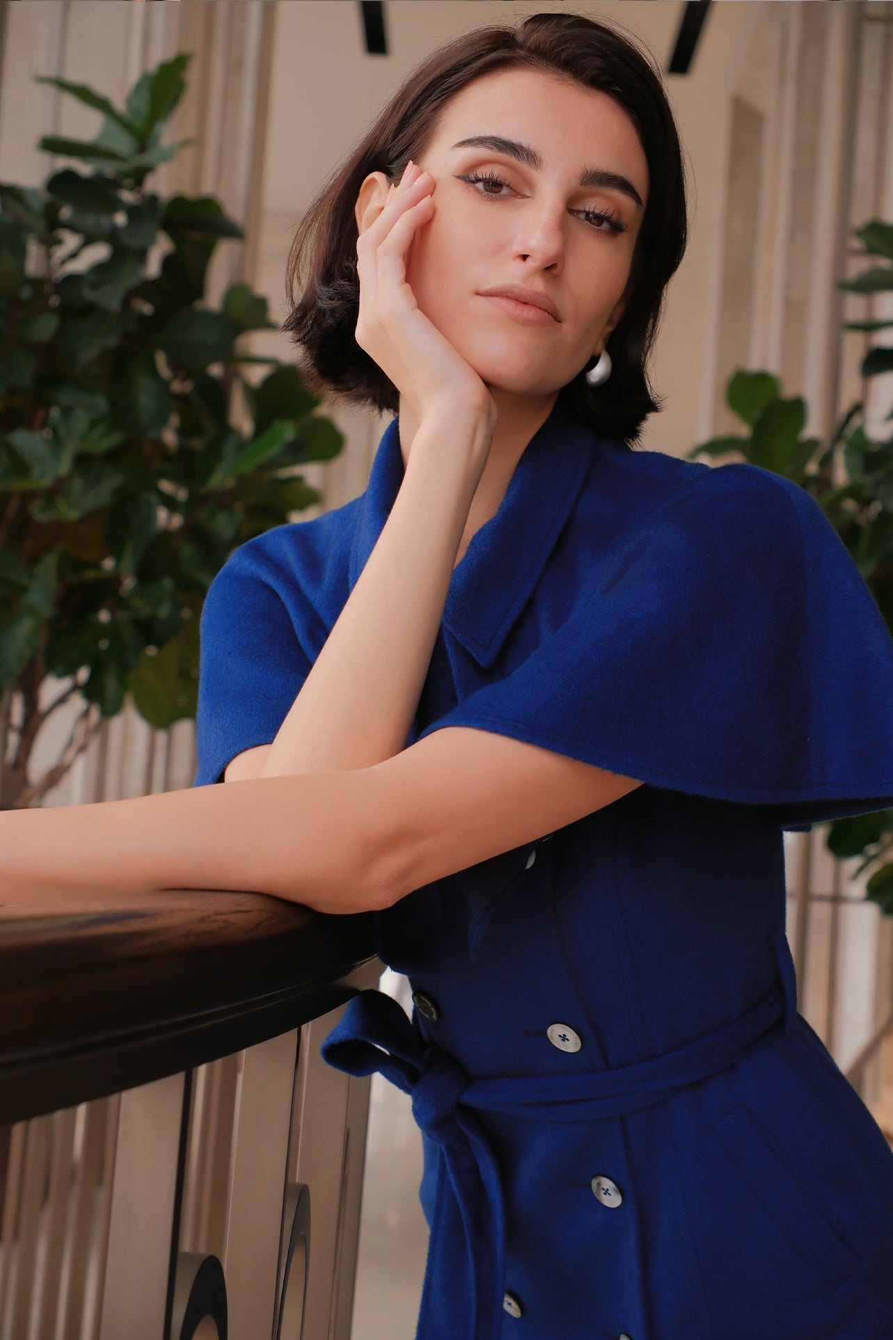 An elegant woman wearing a sophisticated royal cobalt blue trench dress featuring a structured turn-down collar, a cropped capelet overlay, a double-breasted button front, and a cinched self-tie belted waist.