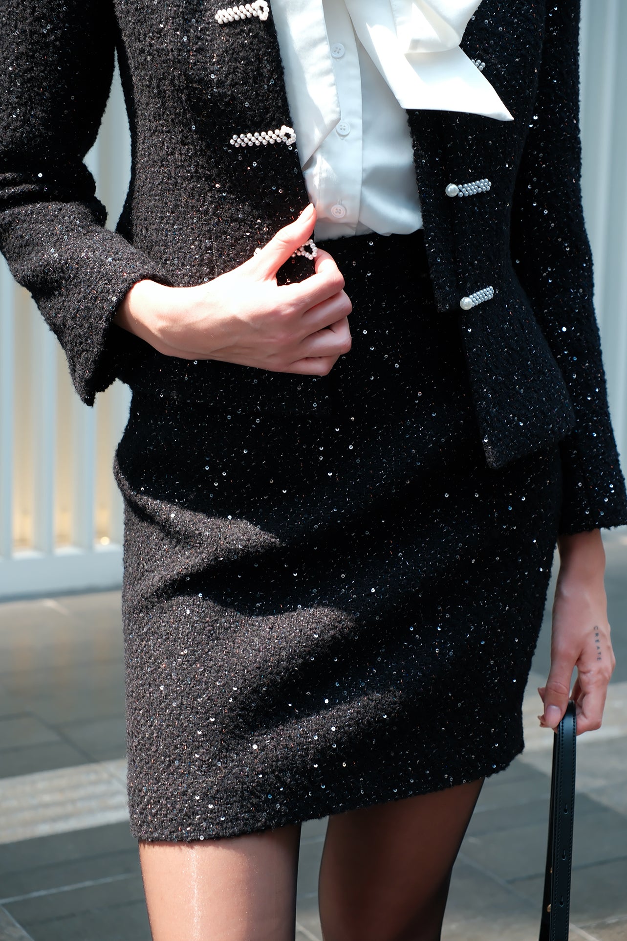 A close-up of a sophisticated woman wearing a high-waisted black sparkle tweed mini pencil skirt featuring subtle glittering sequin accents, styled as part of a matching luxury skirt suit set.