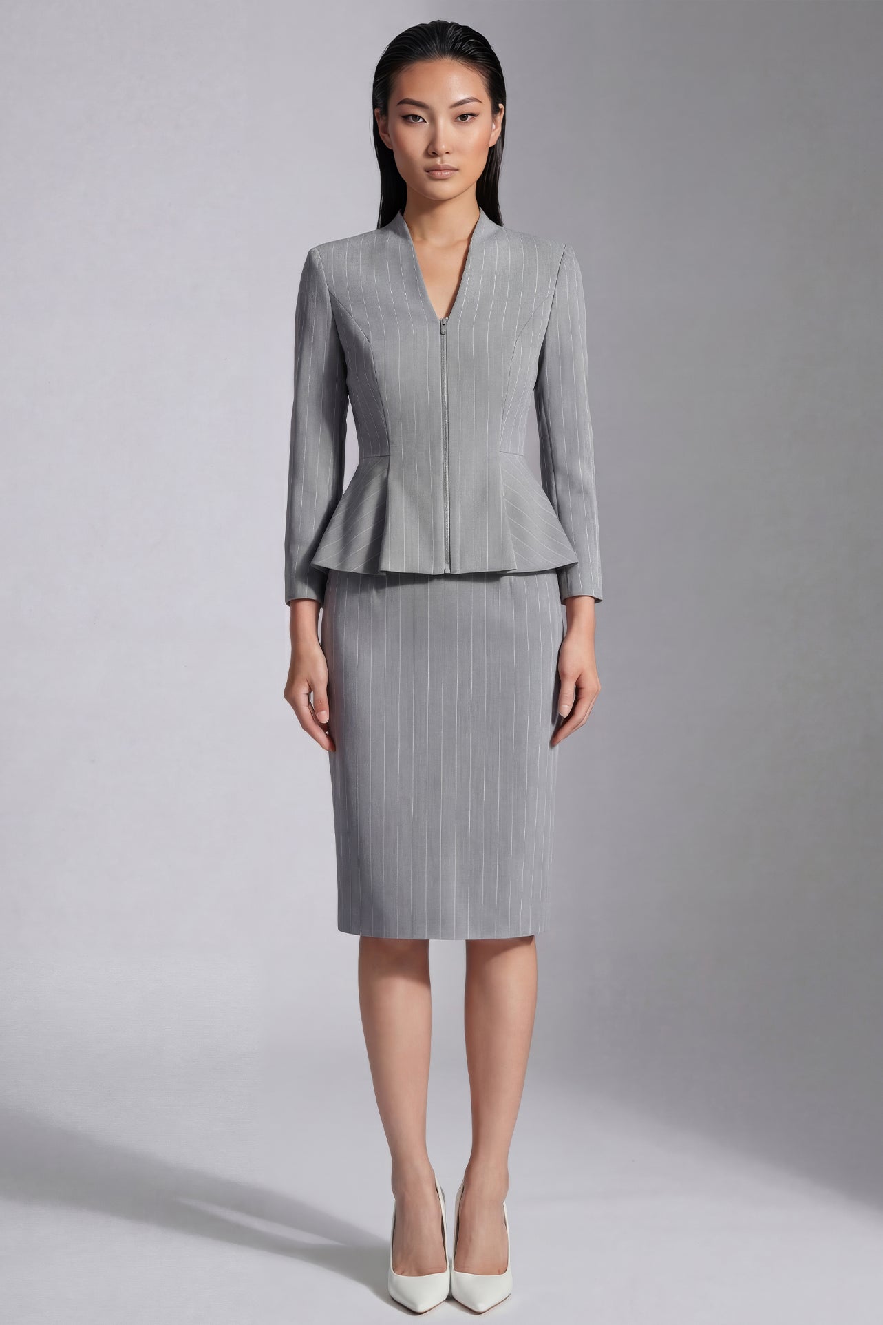 A professional woman wearing a sophisticated grey pinstripe skirt suit, featuring a tailored zipper-front peplum jacket and a matching midi pencil skirt for a polished executive office look.