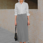 A stylish woman wearing a sophisticated grey pinstripe high-waisted midi skirt with a front slit, paired with a white button-up shirt rolled at the sleeves and black ankle boots for a minimalist corporate chic look