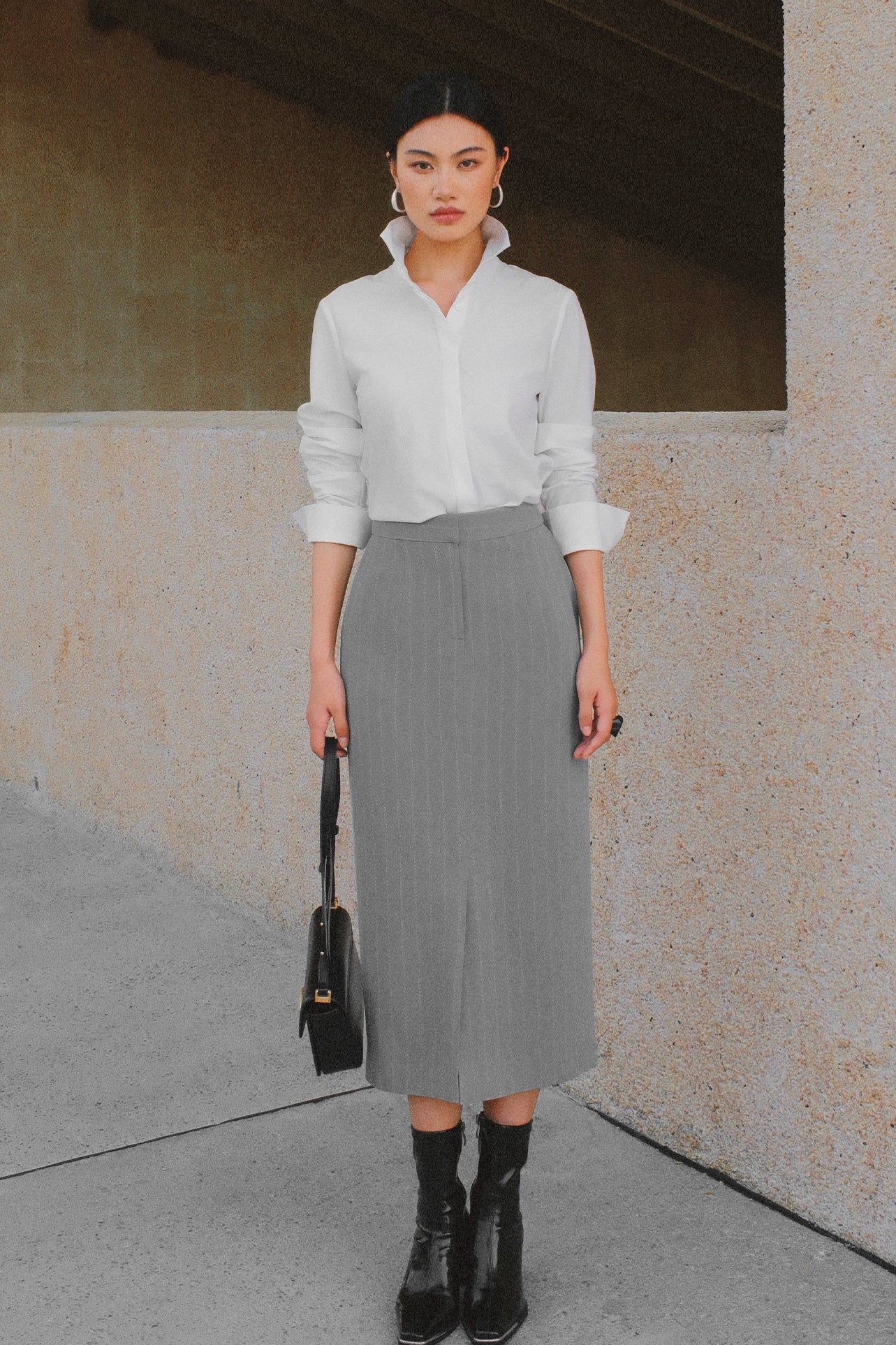 A stylish woman wearing a sophisticated grey pinstripe high-waisted midi skirt with a front slit, paired with a white button-up shirt rolled at the sleeves and black ankle boots for a minimalist corporate chic look