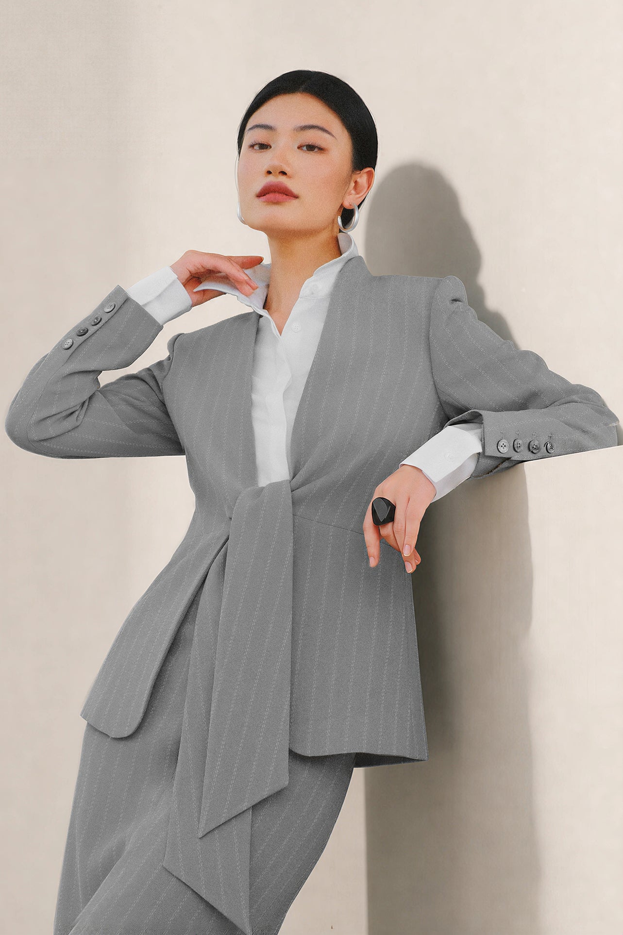 A beautiful professional model wearing an elegant grey pinstripe collarless blazer featuring a distinctive tie-front knot at the waist, styled with a crisp white collared shirt underneath for a modern corporate chic look.