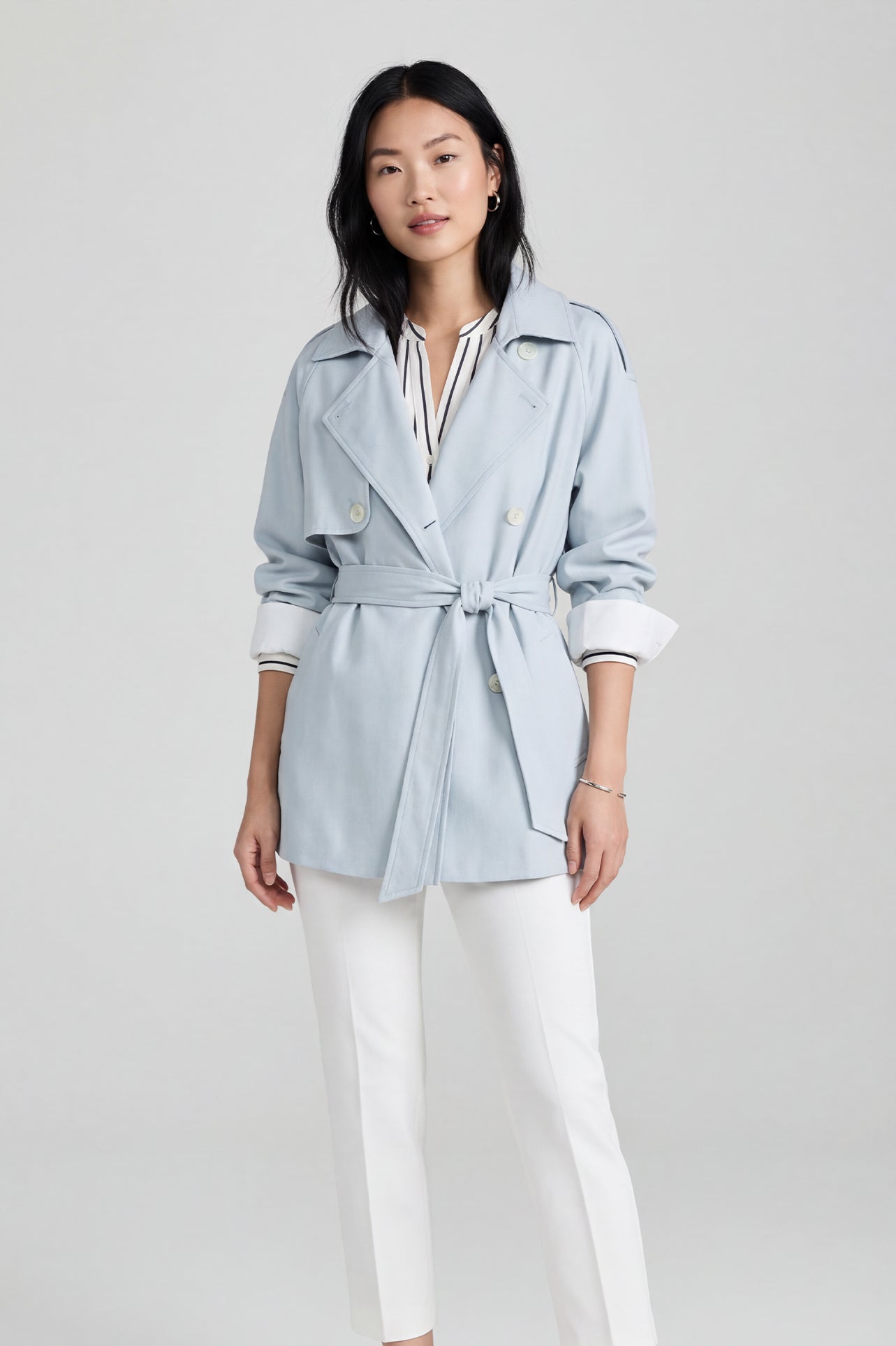 A professional woman wearing an elegant light blue short trench coat with a waist-tie belt and white sleeve cuffs, perfect for a smart casual spring office look.