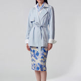 A professional woman wearing an elegant light blue short trench coat with a waist-tie belt and white sleeve cuffs, perfect for a smart casual spring office look.