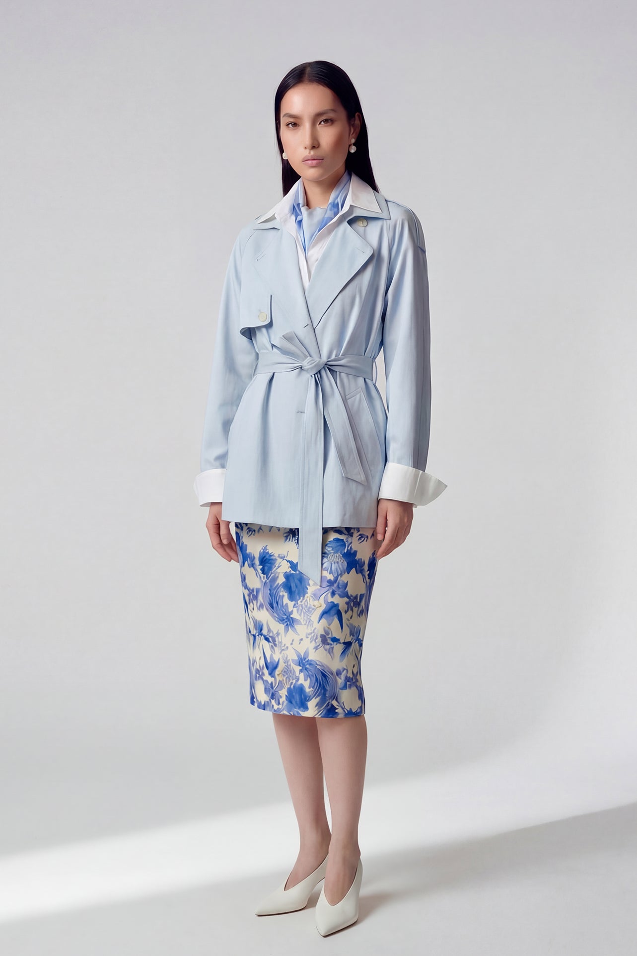 A professional woman wearing an elegant light blue short trench coat with a waist-tie belt and white sleeve cuffs, perfect for a smart casual spring office look.