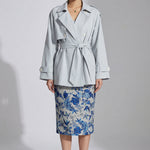 A professional woman wearing an elegant light blue short trench coat with a waist-tie belt and white sleeve cuffs, perfect for a smart casual spring office look.