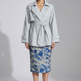 A professional woman wearing an elegant light blue short trench coat with a waist-tie belt and white sleeve cuffs, perfect for a smart casual spring office look.