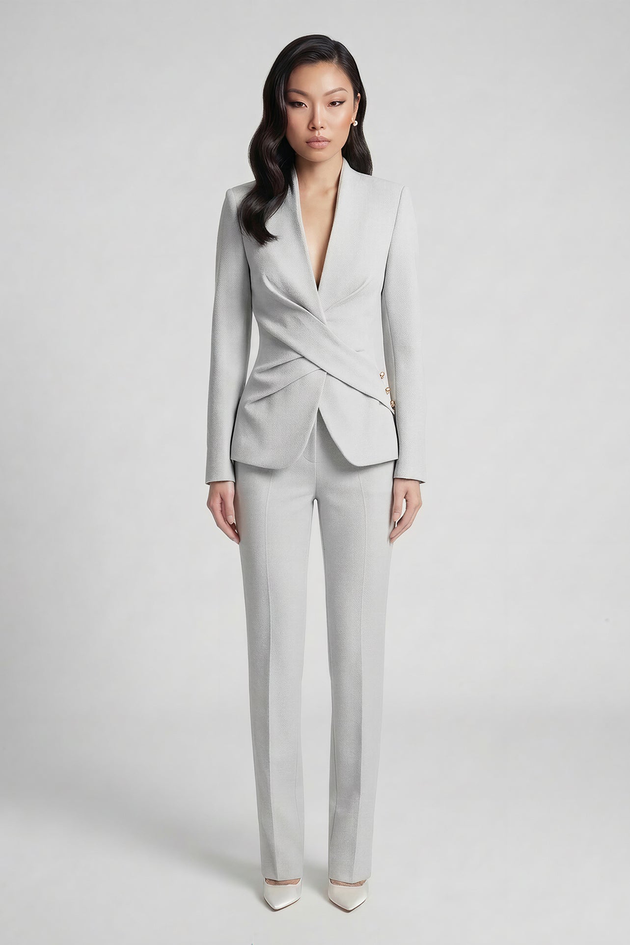 A professional woman wearing a sophisticated light grey tailored suit set, featuring a unique wrap-front blazer with criss-cross waist detailing and matching slim-fit trousers for a polished executive look.