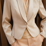 A professional woman modeling a luxurious champagne satin blazer featuring a soft shawl lapel and a single-button closure, elegantly paired with high-waisted white trousers for a sophisticated quiet luxury look.