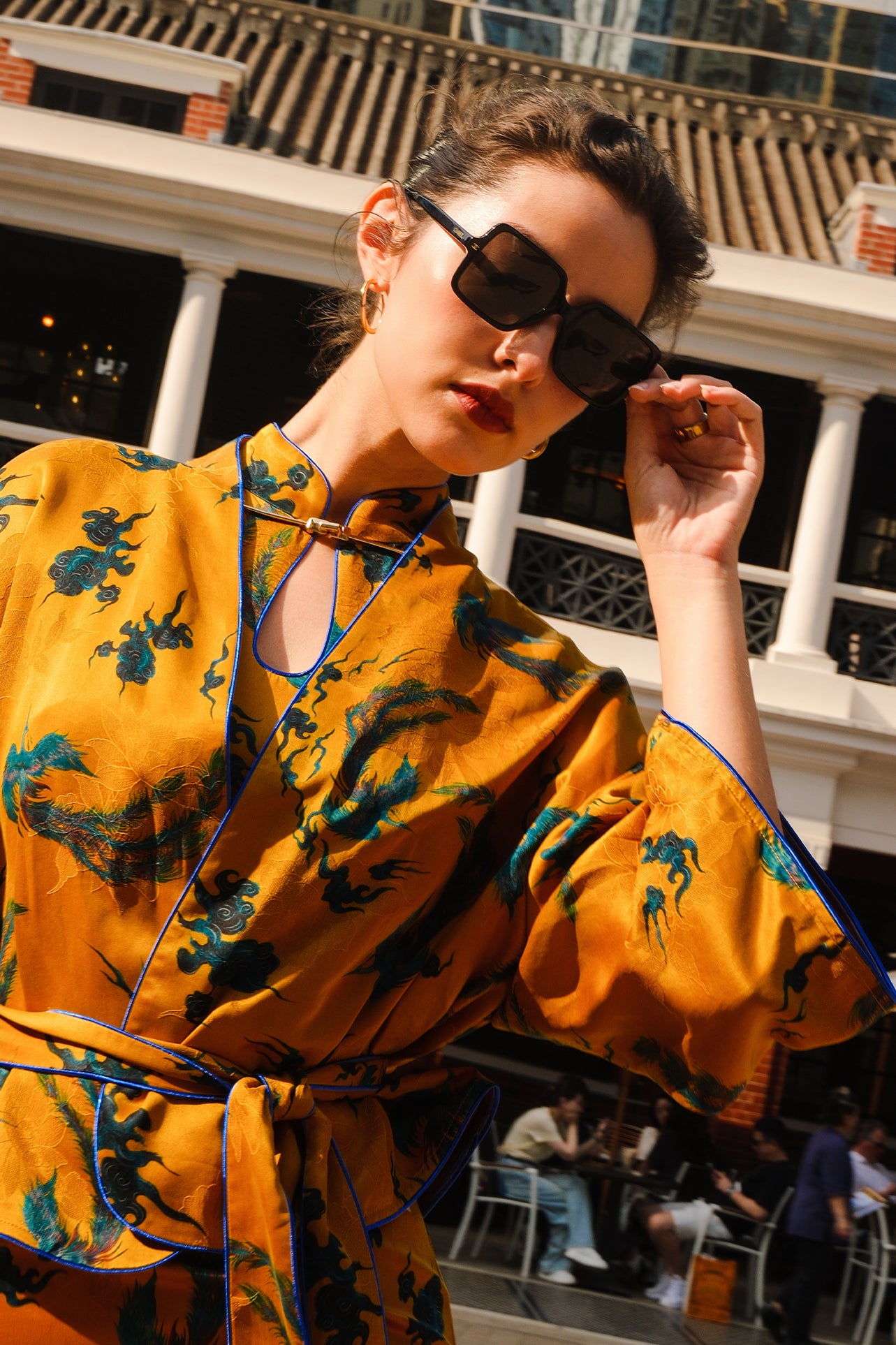 A beautiful woman modeling an elegant mustard yellow modern Chinese jacket, featuring intricate blue phoenix embroidery, a sophisticated mandarin collar, and wide kimono sleeves with a matching waist tie