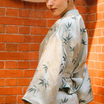 A professional woman modeling an elegant light sage green silk wrap jacket, featuring dark green bamboo leaf prints, a sophisticated mandarin collar with keyhole detail, and wide kimono sleeves.