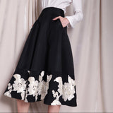 An elegant woman wearing a sophisticated black A-line midi dress, featuring exquisite white floral embroidery along the hem and professional three-quarter sleeves for a timeless formal look.