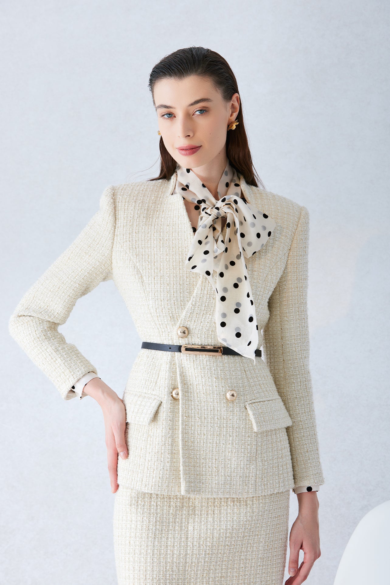 A professional woman wearing a sophisticated cream white tweed skirt suit, featuring a double-breasted blazer with gold buttons, a slim black belt, and an elegant polka dot pussy bow tie for a polished French chic office look.