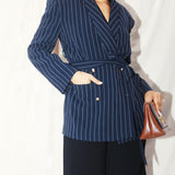 A professional woman wearing a sophisticated navy blue pinstripe blazer jacket, featuring a longline silhouette with a matching tie-waist belt and gold earring accents for an elegant executive office look.