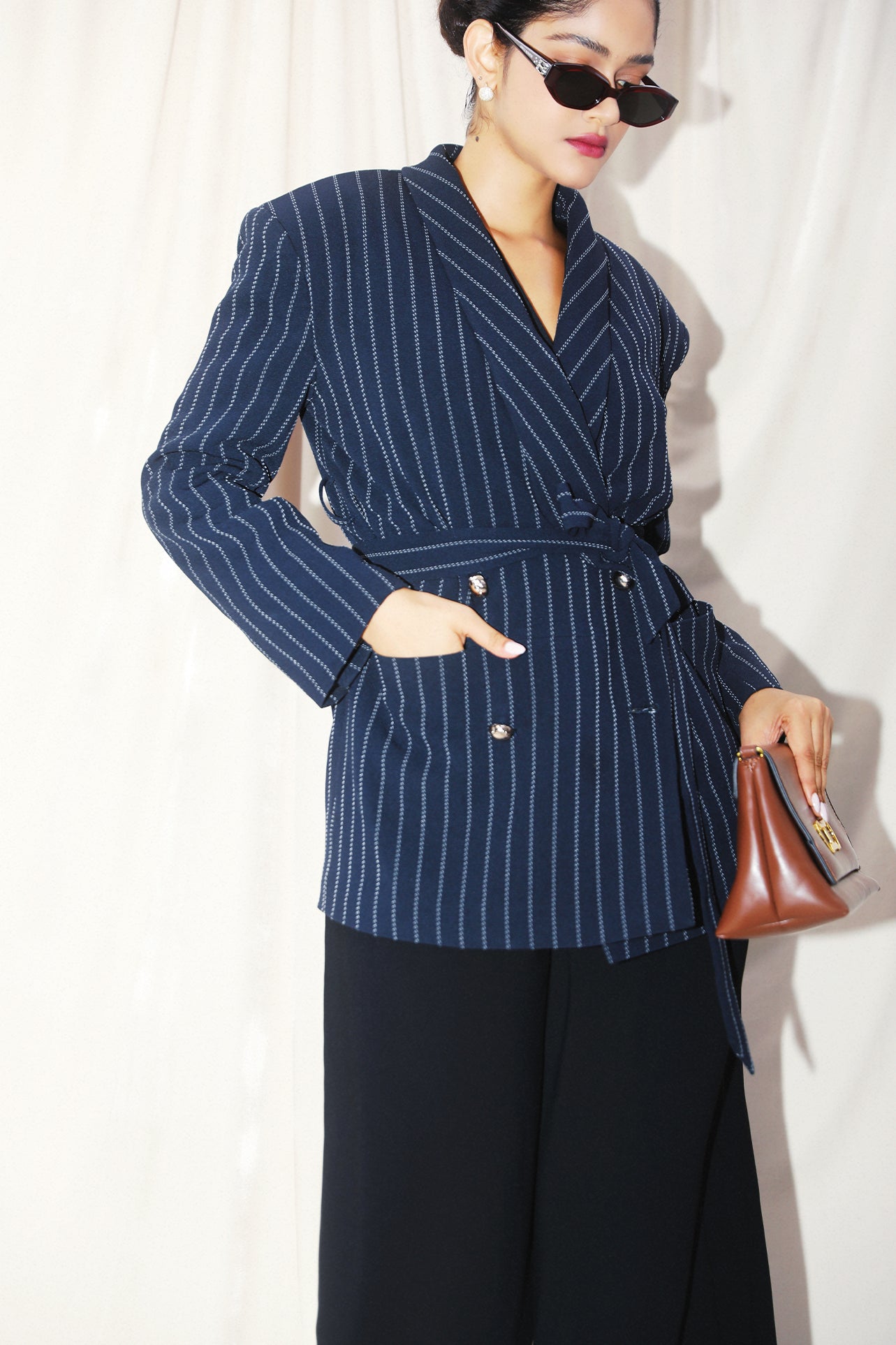 A professional woman wearing a sophisticated navy blue pinstripe blazer jacket, featuring a longline silhouette with a matching tie-waist belt and gold earring accents for an elegant executive office look.