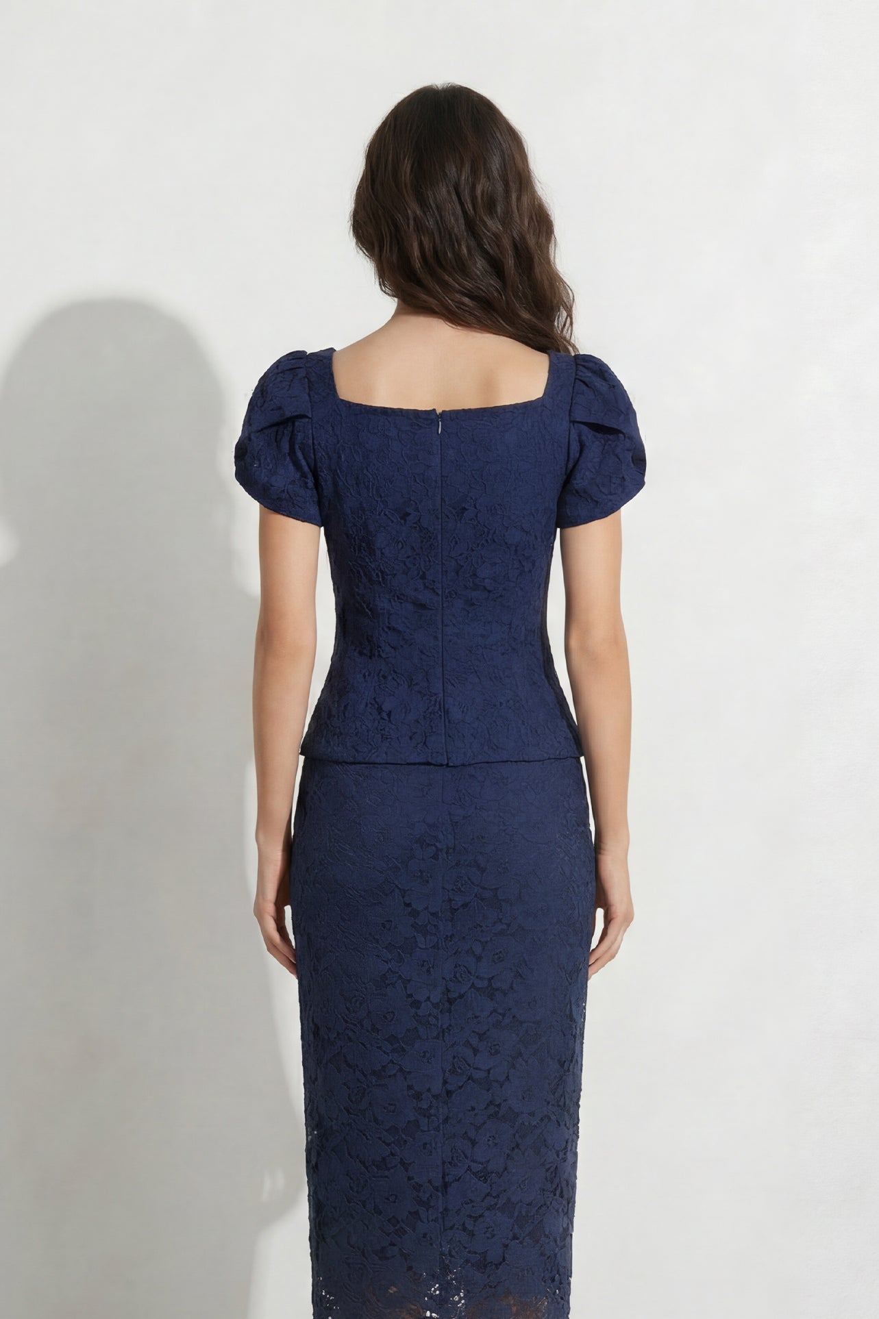 A professional woman modeling an elegant navy blue lace short top, featuring a retro square neckline, romantic short puff sleeves, and a series of pearl and rhinestone frog closures on the front with a tailored triangular split hem, paired with light blue high-waisted jeans.