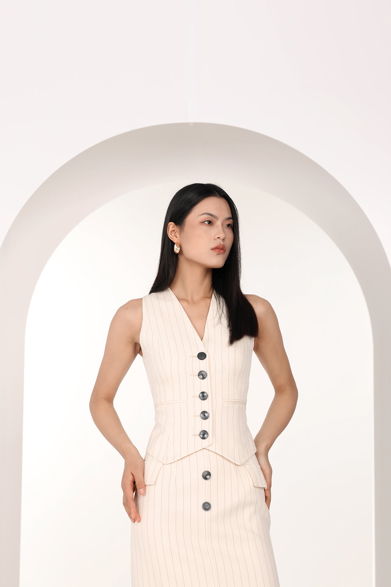 A professional woman wearing a sophisticated off-white pinstripe vest top, featuring a slim-fit tailored silhouette, classic V-neck, and contrasting black buttons for a modern business casual look.