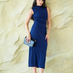 A professional woman wearing a sophisticated navy blue sleeveless midi dress, featuring an elegant high neckline and flattering ruched detailing along the side for a slim-fit executive look.
