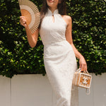 A professional woman modeling an elegant white lace halter neck modern qipao dress, featuring a sophisticated mandarin collar with keyhole detail and an intricate eyelet embroidered fabric for a quiet luxury look.