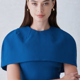 A professional woman wearing a sophisticated teal blue midi dress with an elegant cape-style overlay and structured shoulders, perfect for formal executive events and ceremonial occasions.