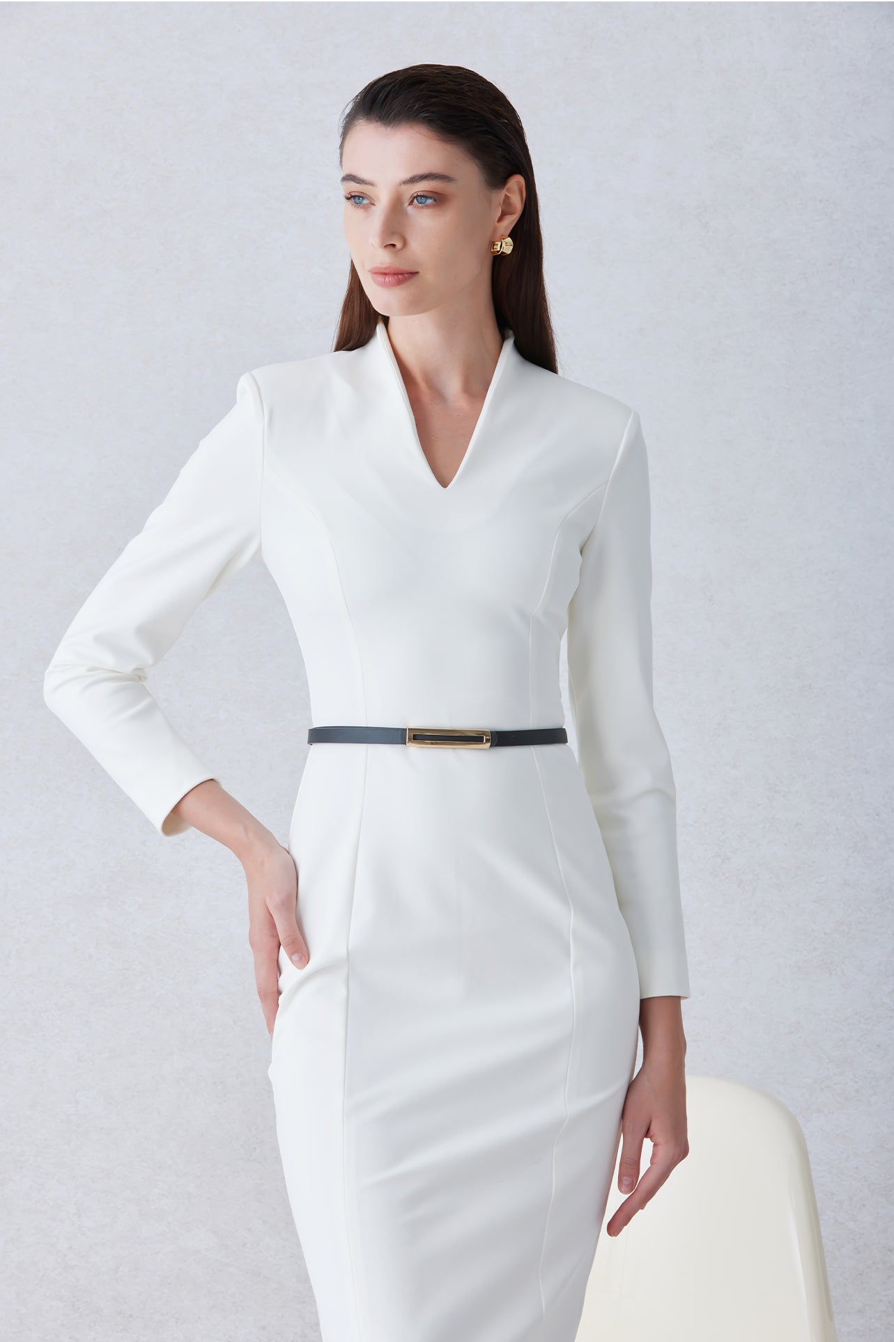 A professional woman wearing a sophisticated solid white long-sleeve sheath dress, featuring an elegant V-neckline and a slim black belt with a gold buckle for a polished executive office look.