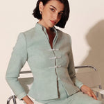 An elegant woman sitting on a modern chair wearing a sophisticated mint green textured suit featuring a cropped jacket with a mandarin collar, three white frog closures, and a matching high-waisted front-slit pencil skirt.