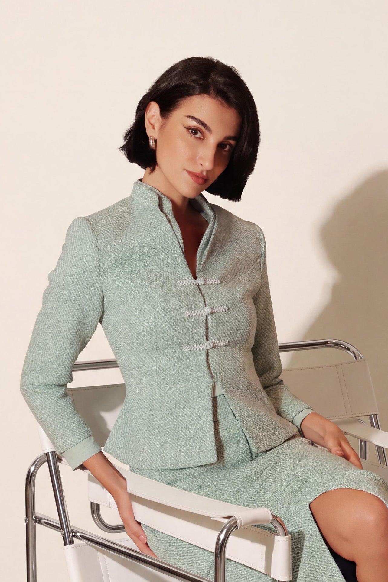 An elegant woman sitting on a modern chair wearing a sophisticated mint green textured suit featuring a cropped jacket with a mandarin collar, three white frog closures, and a matching high-waisted front-slit pencil skirt.