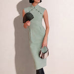A stylish woman wearing sunglasses and holding a black folding fan, dressed in an elegant mint green sleeveless modern qipao dress featuring a mandarin collar with delicate button trim along the bodice and a tailored midi sheath silhouette.
