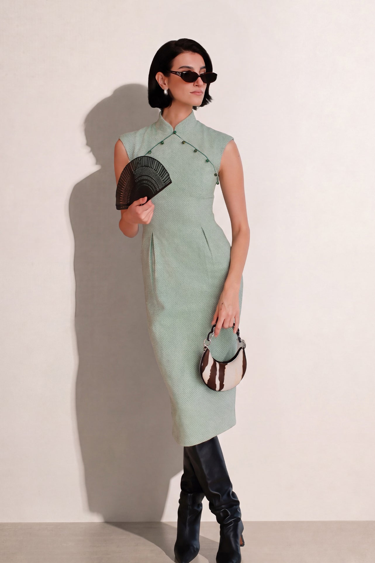 A stylish woman wearing sunglasses and holding a black folding fan, dressed in an elegant mint green sleeveless modern qipao dress featuring a mandarin collar with delicate button trim along the bodice and a tailored midi sheath silhouette.