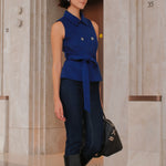 A style comparison of two professional women wearing modern cobalt blue outerwear featuring self-tie waist belts; on the left, a tailored cropped capelet jacket paired with jeans, and on the right, a double-breasted sleeveless vest with white contrast buttons styled with black trousers