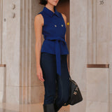 A style comparison of two professional women wearing modern cobalt blue outerwear featuring self-tie waist belts; on the left, a tailored cropped capelet jacket paired with jeans, and on the right, a double-breasted sleeveless vest with white contrast buttons styled with black trousers