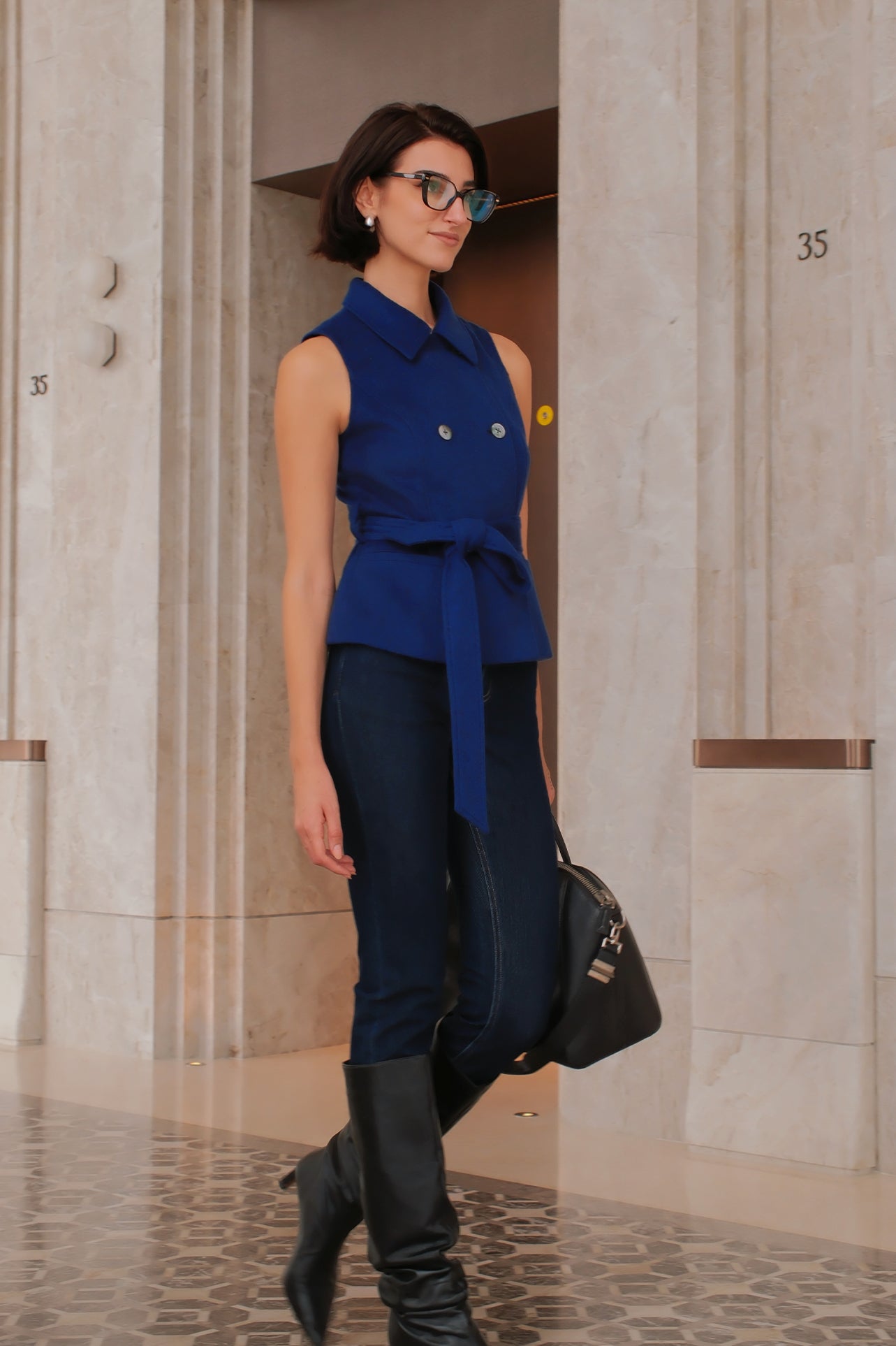 A style comparison of two professional women wearing modern cobalt blue outerwear featuring self-tie waist belts; on the left, a tailored cropped capelet jacket paired with jeans, and on the right, a double-breasted sleeveless vest with white contrast buttons styled with black trousers